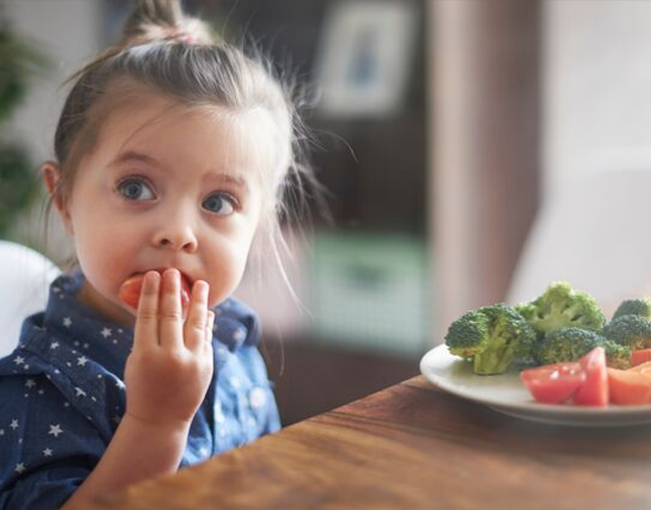 Little girl eating a tomato next to a table with a plate filled with broccoli and tomatoes